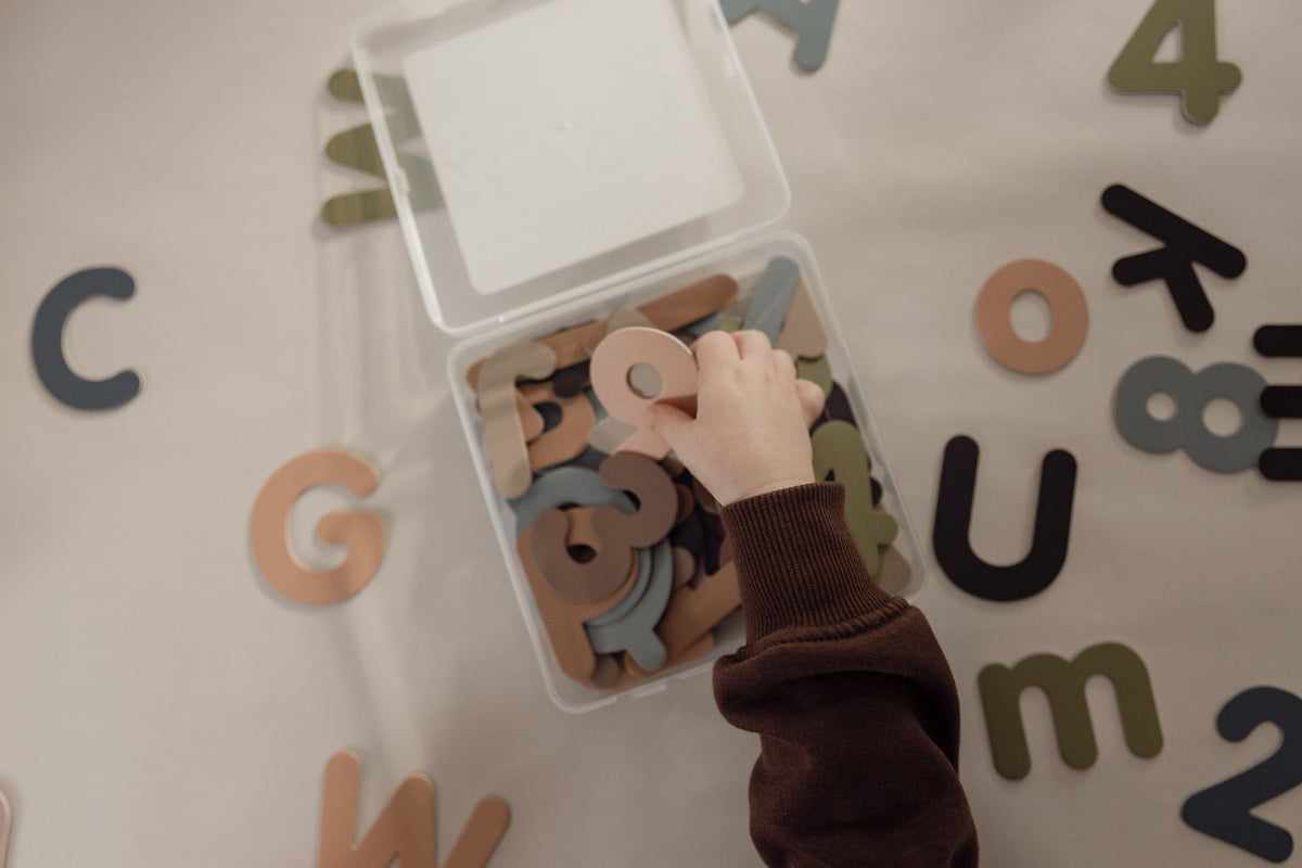 Child's hand reaching into a clear box of magnetic letters and numbers in soft, muted colours