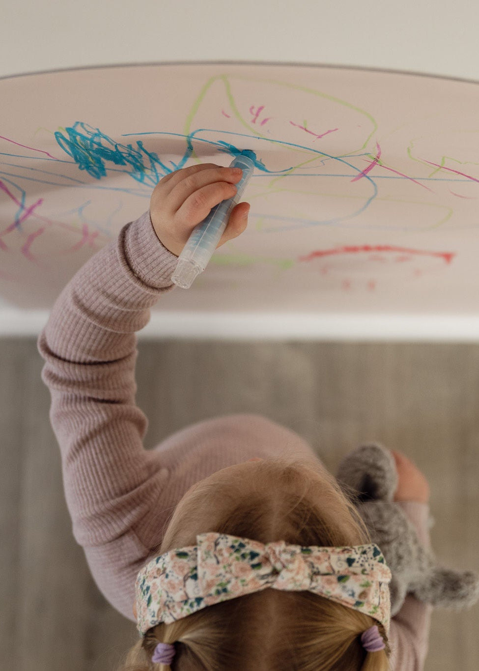 A child wearing a floral patterned headband and a mauve ribbed sweater draws on a large light blush magnetic arch decal. The child holds blue chalk, and the scene is shot from a high-angle perspective above against a grey wood-look floor.