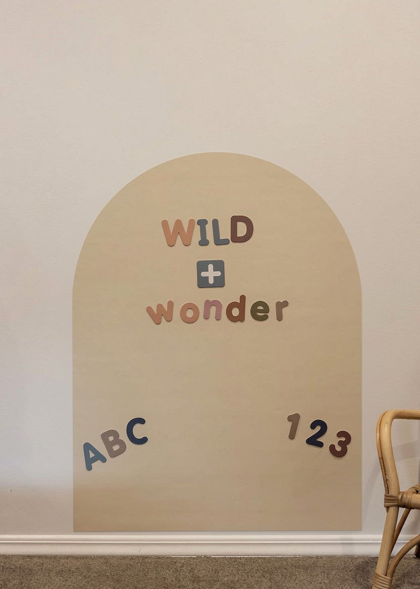 A beige magnetic arched wall decal with the words “WILD + wonder” and small letters/numbers placed on it, shown against a light wall above a carpeted floor with part of a wooden chair visible at the right edge.