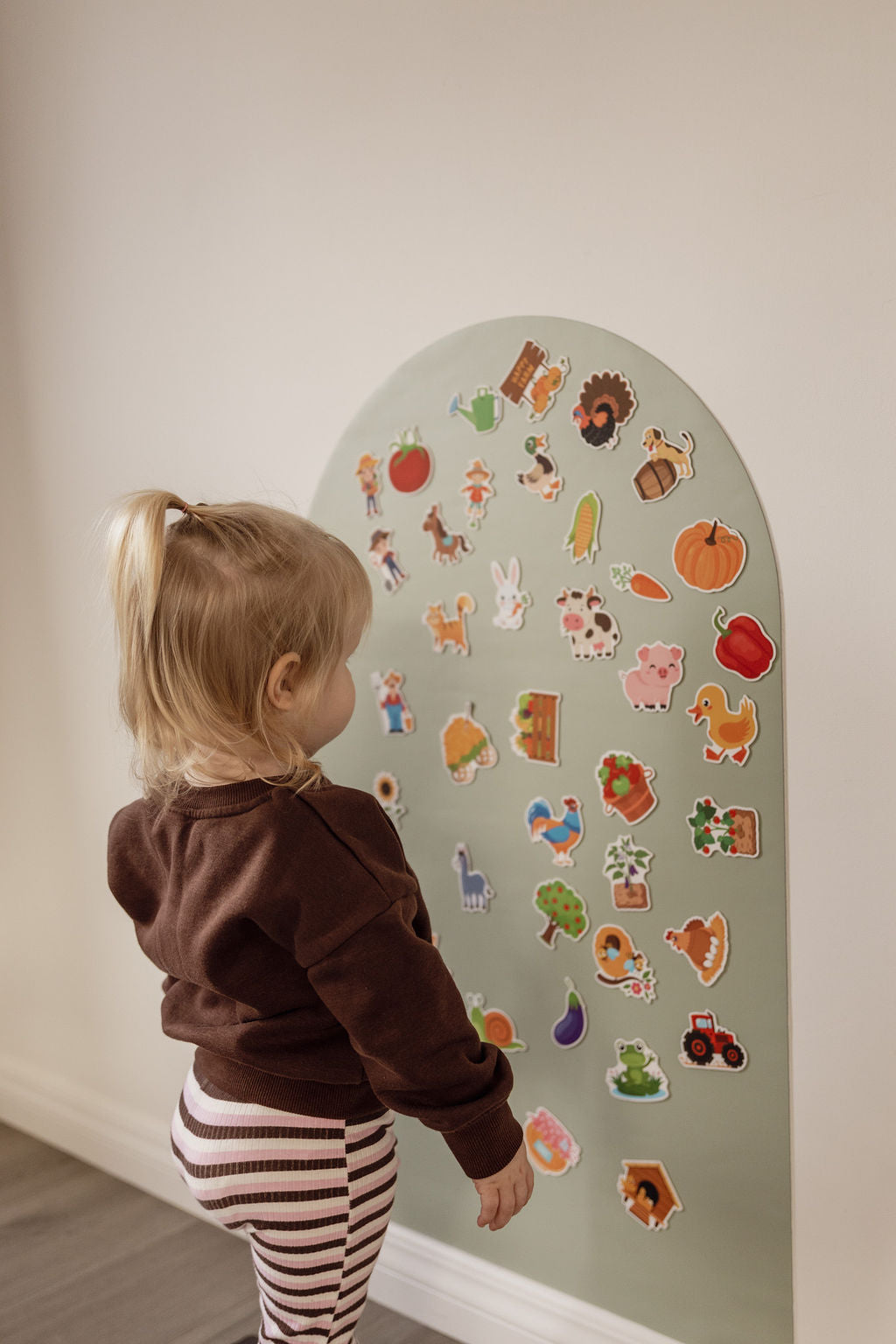 A young child with blonde hair in a ponytail stands in profile facing a large arched magnetic wall decal covered in colourful farm animals, fruits, and vegetables. The child is wearing a dark brown sweatshirt and brown-and-white striped pants, set against a neutral beige wall and light wood floor.