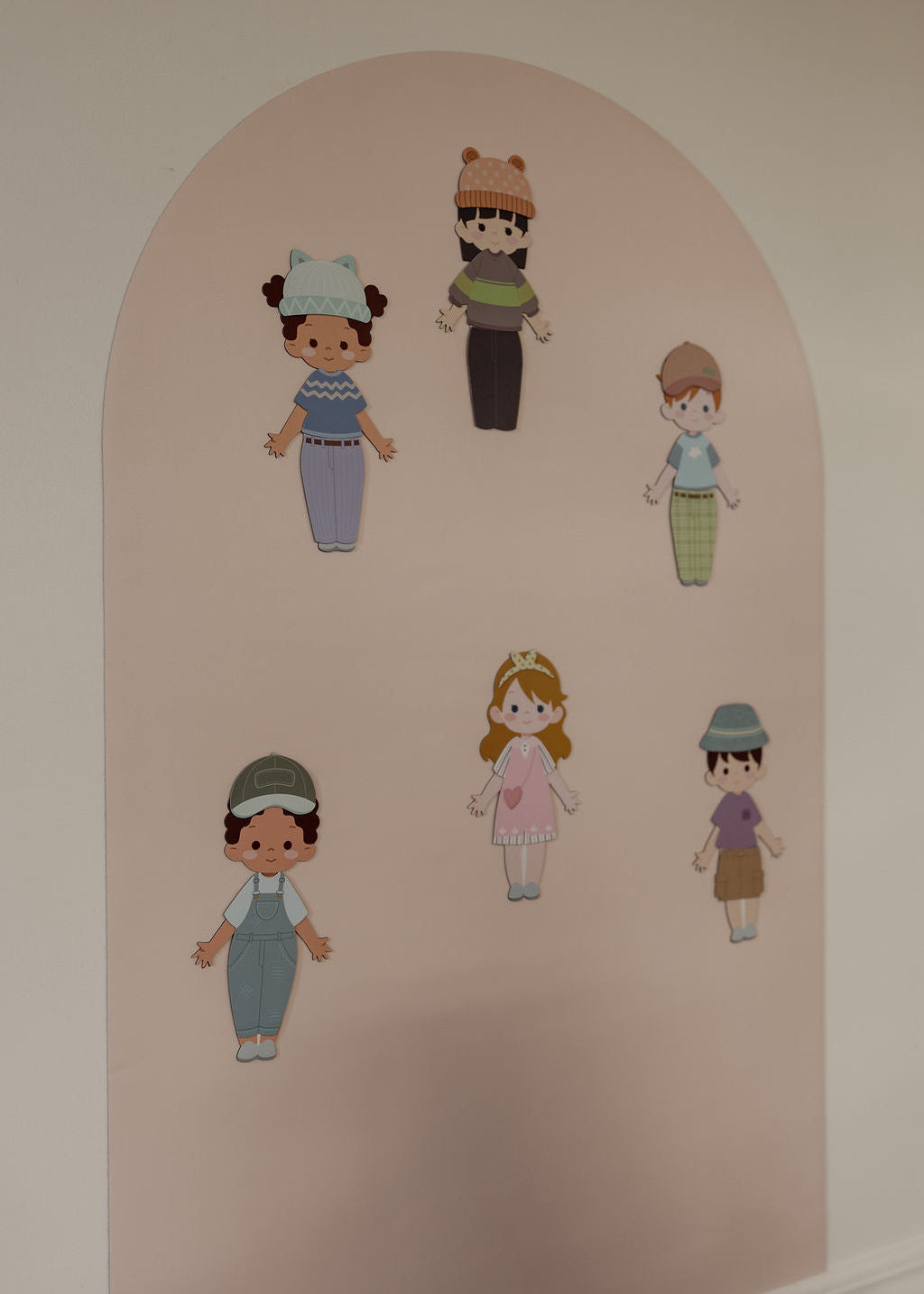A set of six magnetic cutout children's characters in various outfits, arranged on a light pink magnetic arched wall decal against a white wall.