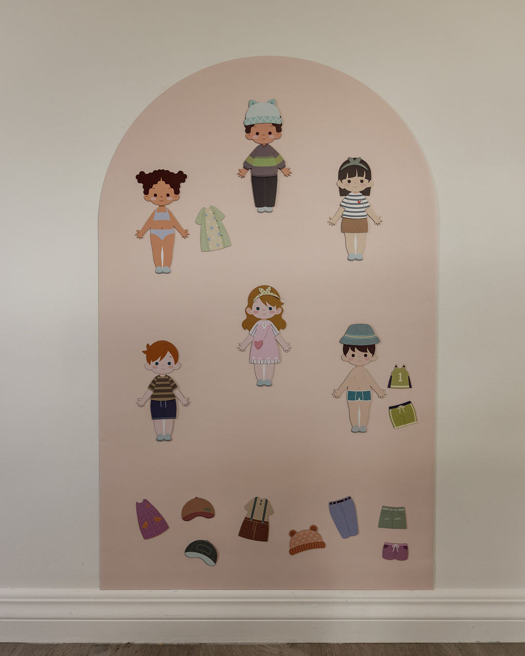 A children's wall-mounted magnetic arch decal featuring illustrated cut out doll characters and small clothing pieces on a pale pink arched panel.