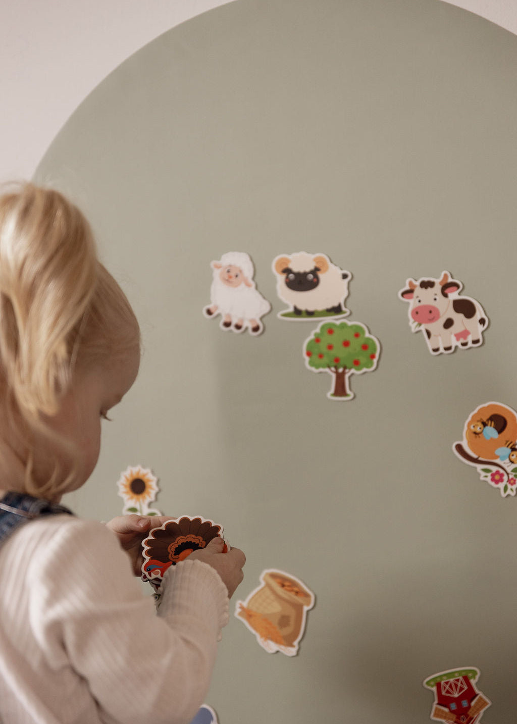 A young child with blonde hair is seen from behind, interacting with colourful  farm animal magnets on a large, light sage green magnetic wall arch decal.