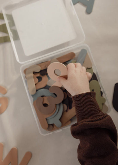 A child's hand reaches into a clear plastic storage box filled with magnetic alphabet letters, with more scattered around on a light beige surface.